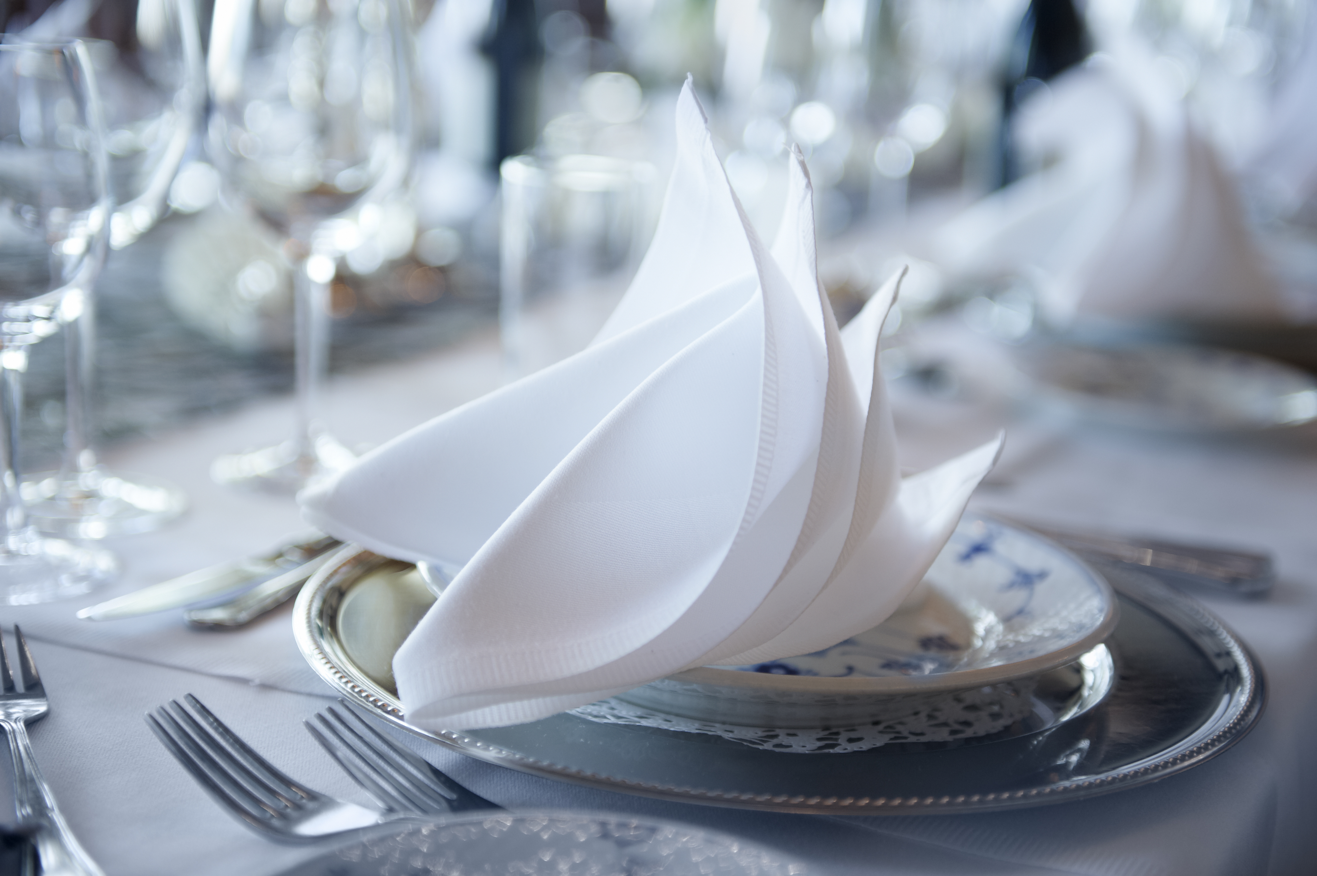 An origami napkin on a formal restaurant plate, with forks in the foreground and glasses in the background